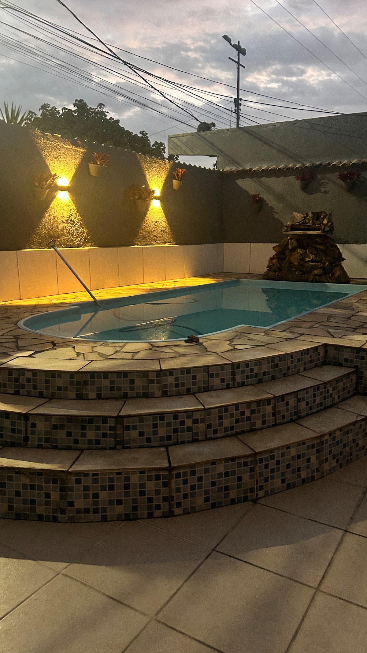 The outdoor area features a crystal-clear pool surrounded by stone steps. Soft lighting highlights the textured walls and decorative plants, creating a relaxing ambiance. A stone feature resembling a waterfall is positioned nearby, enhancing the tranquil feel of the space.
