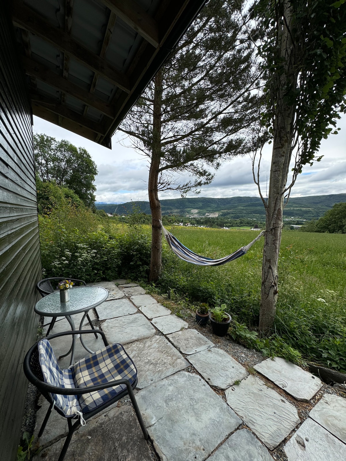 A small outdoor patio area is visible, featuring a round glass table and two checkered chairs. A hammock is strung between two trees, set against a backdrop of lush greenery and rolling hills. The sky is partly cloudy, adding to the serene atmosphere.