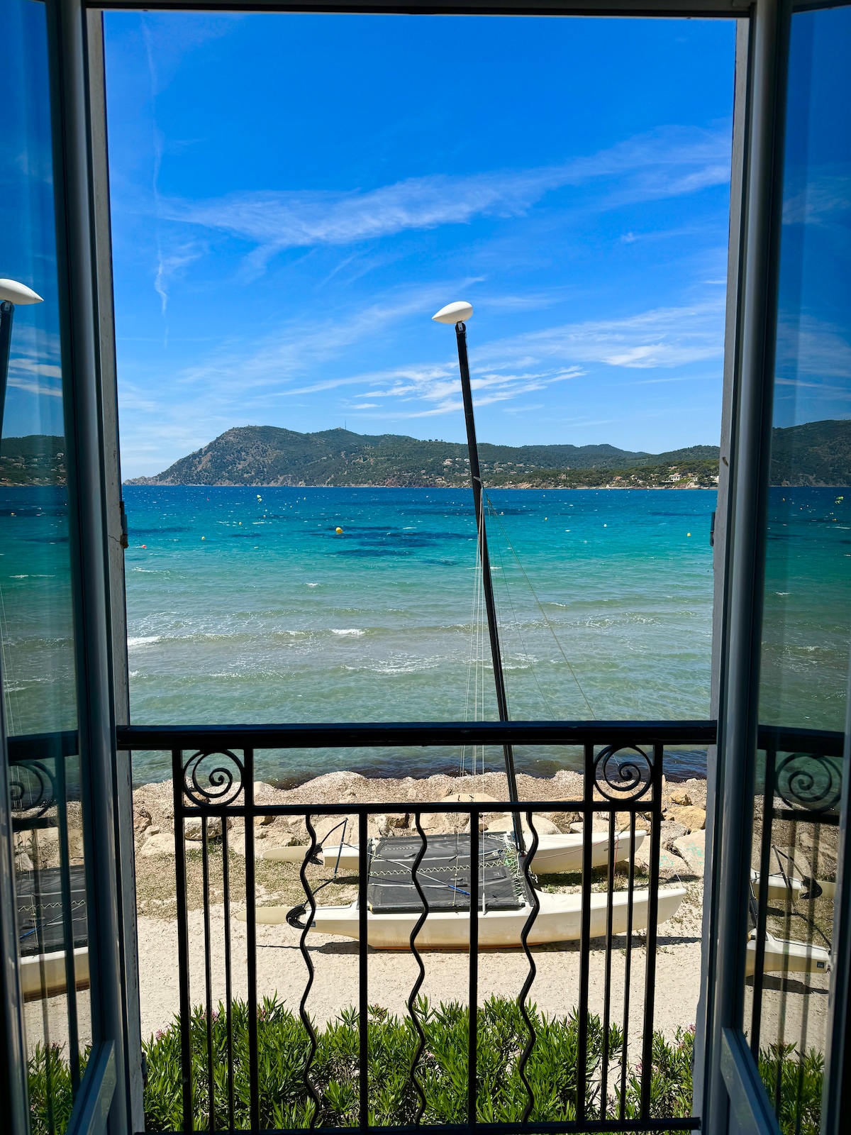 A scenic view of the sea is framed by large, open French doors, revealing sparkling blue waters and a distant mountainous shoreline. The foreground features a rocky beach area, enhancing the coastal atmosphere. A gentle breeze is suggested by the movement of the water.