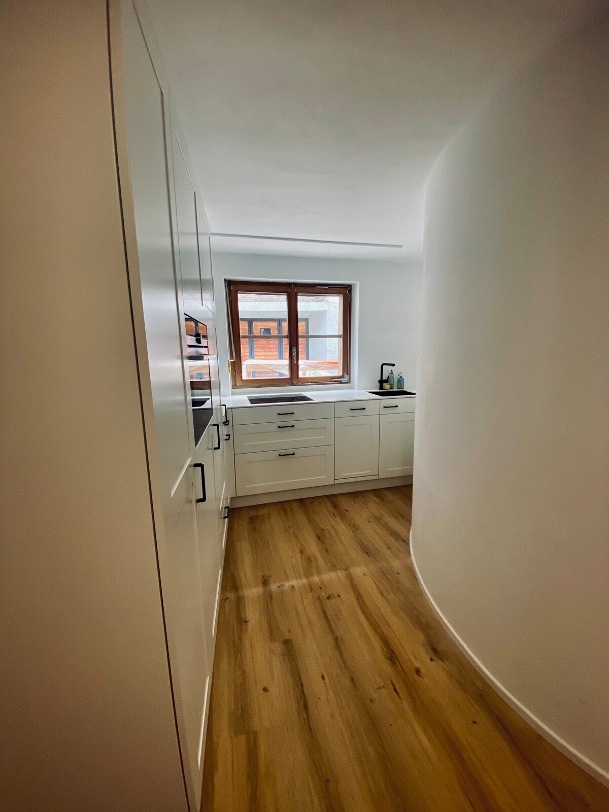A compact kitchen is viewed from a hallway, featuring modern cabinetry and a countertop. Natural light filters in through a window, reflecting against the light-colored walls and wooden floor. The curved wall adds a unique architectural element to the space.