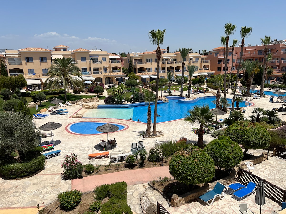 A large communal swimming pool is surrounded by landscaped gardens and flower beds. Palm trees and umbrellas provide shaded seating areas. Lounge chairs are arranged around the pool, and the buildings of the complex can be seen in the background against a clear blue sky.