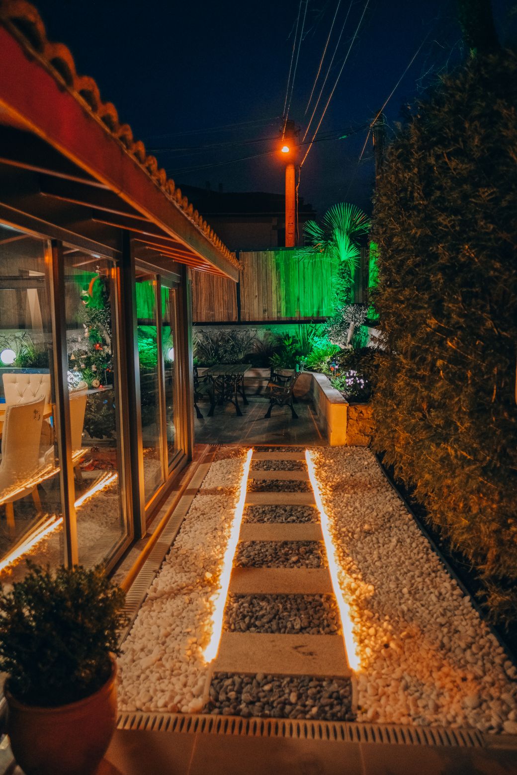 A pathway lined with illuminated stones leads to a seating area surrounded by greenery. Soft lighting from the garden highlights the space, while glass walls reflect a cozy atmosphere in the evening. The outdoor area is designed for relaxation and enjoyment.