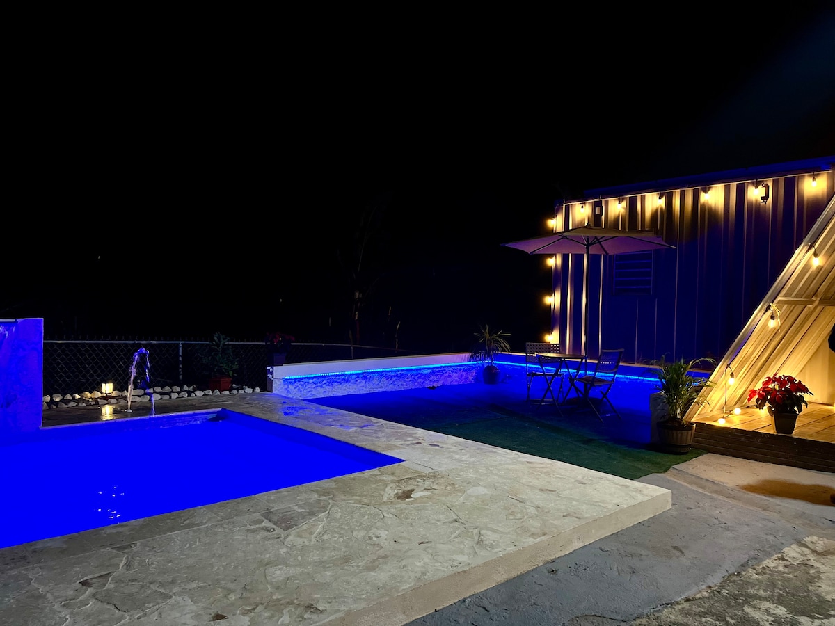 A swimming pool is illuminated with blue lighting, reflecting off the surrounding stone surface. Ambient lighting is strung along a nearby building, with a patio area featuring a table and umbrella. Potted plants add greenery to the inviting outdoor space.