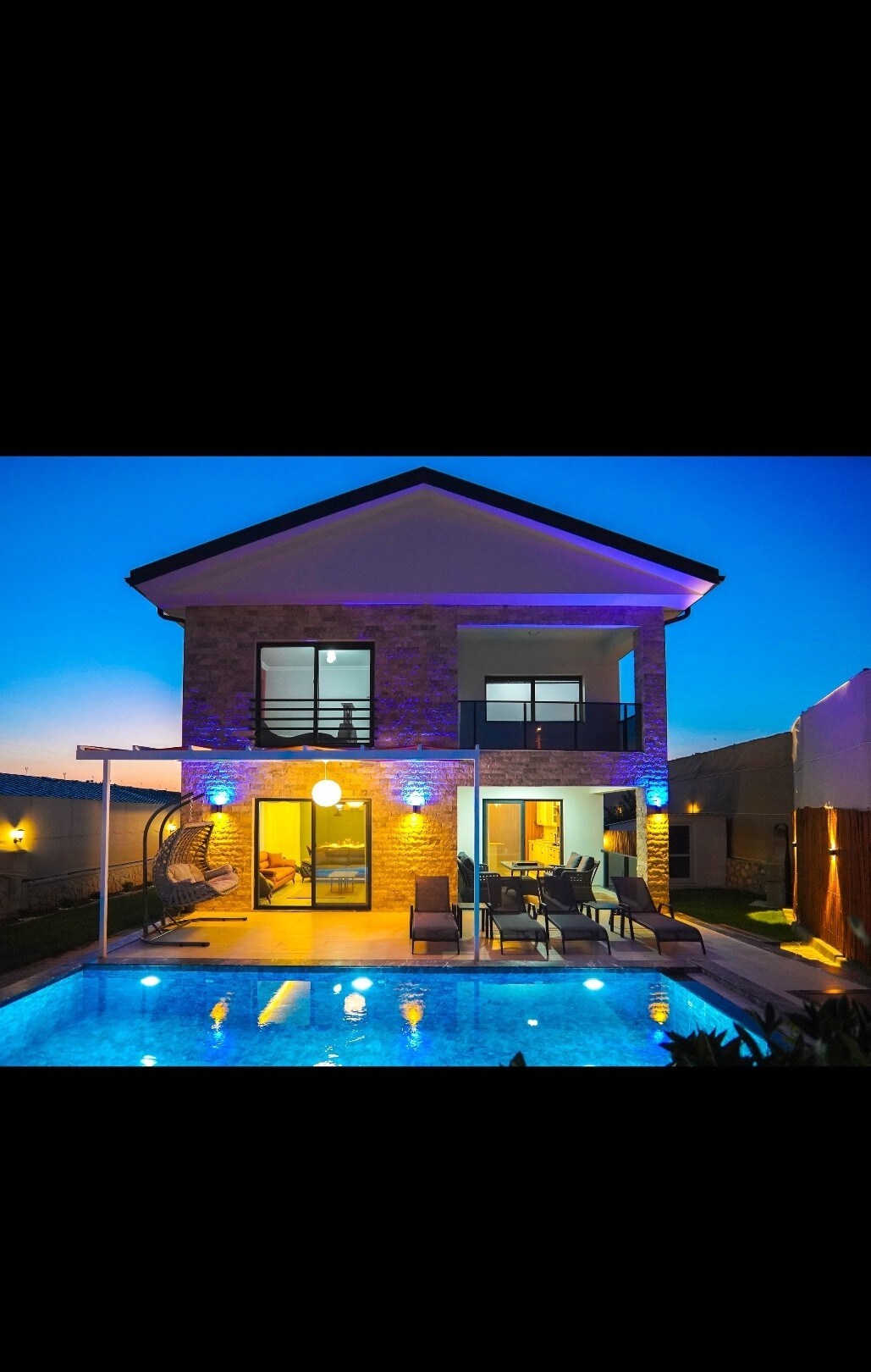 A modern villa is highlighted at dusk, featuring a well-lit facade and a private pool reflecting soft blue tones. Outdoor seating is arranged beside the pool, and large windows offer glimpses into the inviting indoor space.