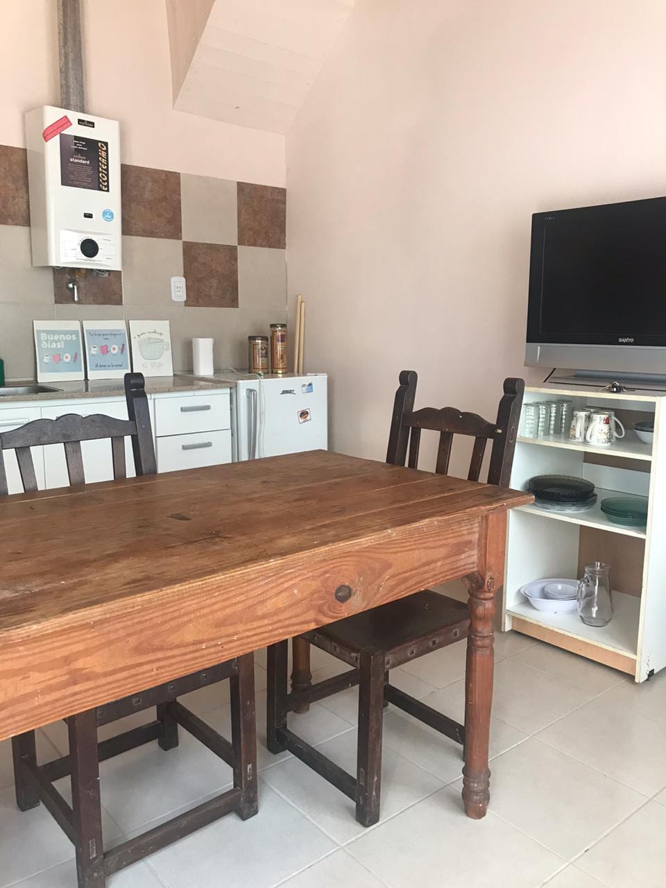 A wooden dining table is set with two chairs, positioned in a light-colored kitchen area. A compact refrigerator and kitchen appliances are neatly arranged nearby. A television sits on a shelf, which also displays various kitchenware, contributing to the functional ambiance.