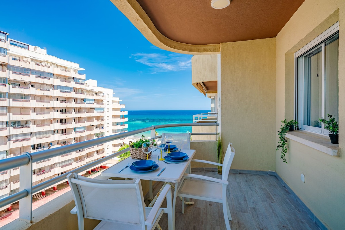 An outdoor terrace is depicted, featuring a dining table set with dishes and cutlery. White chairs are placed around the table. The backdrop showcases a stunning view of the Mediterranean Sea and nearby buildings against a clear blue sky.