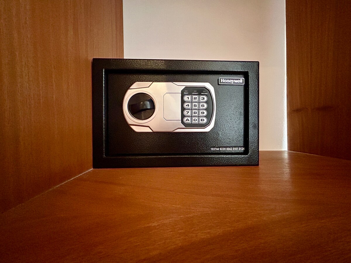 A secure electronic safe is positioned within a wooden cabinet. The safe features a keypad with buttons for numeric input, facilitating access to stored items. The cabinet showcases a smooth wooden surface, complementing the overall design of the space.