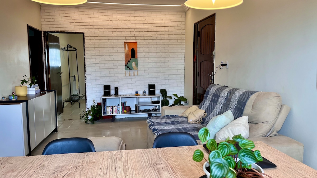 A living area features a beige couch draped with a blue and white blanket. A low media unit displays books and plants. The light-colored walls enhance the space, while a dining table with black chairs sits in the foreground, providing a sense of openness.