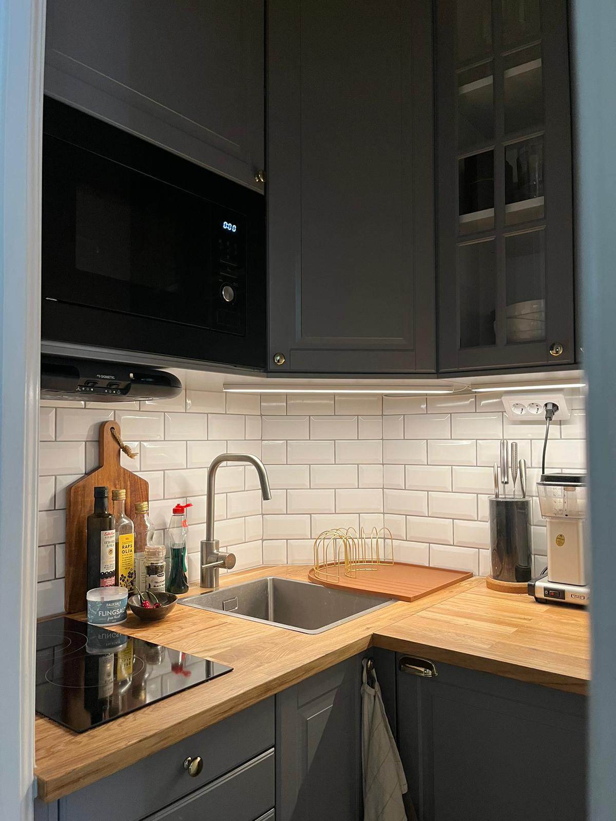 The kitchen features dark cabinets contrasted by a light tile backsplash, with a wooden countertop and integrated sink. A microwave and an induction cooktop are visible, accompanied by essential kitchen tools and condiments arranged neatly on the counter.