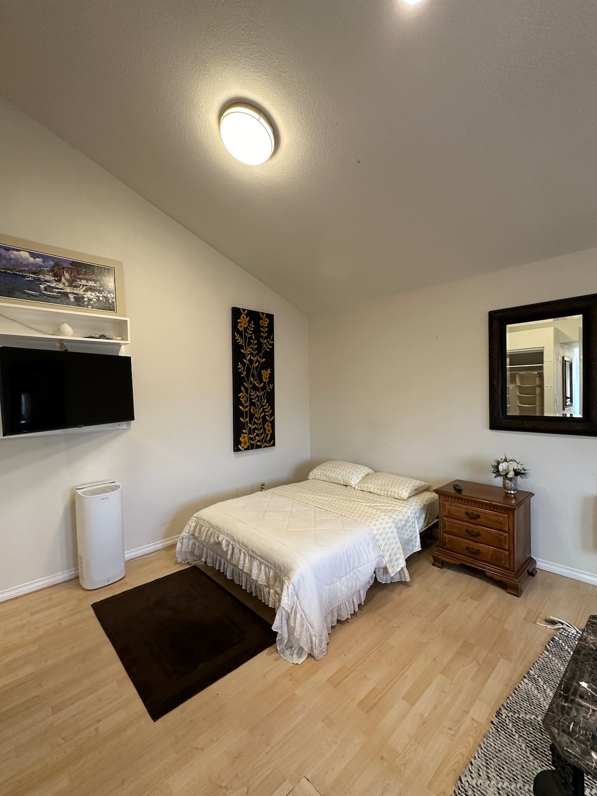 A minimalist bedroom is presented with a spacious bed covered in a light quilt. A small wooden nightstand sits beside the bed, with a decorative flower arrangement. A wall-mounted television and a large mirror enhance the room's functionality, while soft lighting contributes to a calm ambience.