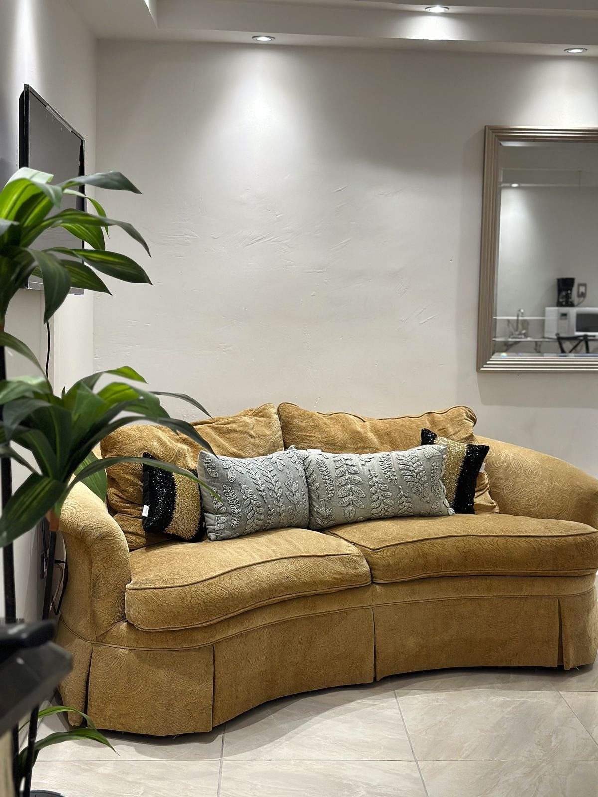 A cozy two-seater couch with a soft, curved design is displayed against a light-colored wall. Two decorative pillows with subtle patterns rest on the couch. A tall green plant adds a natural touch to the space, enhancing the warm atmosphere.
