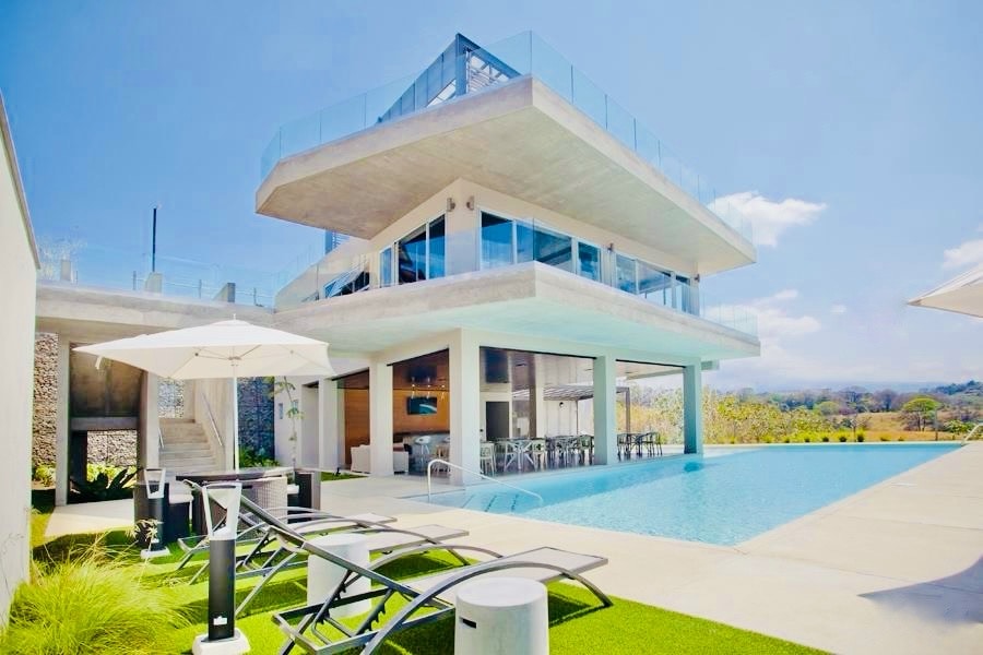 A modern building with multiple levels is highlighted by expansive glass walls and a rooftop terrace. An infinity pool stretches alongside sun loungers on a grassy area, reflecting the clear blue sky and surrounding landscape.