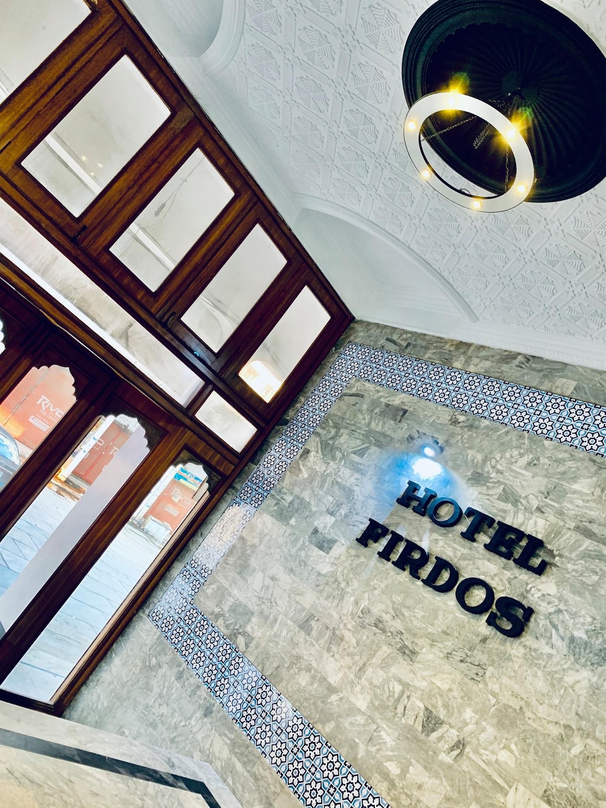 A welcoming lobby features wooden doors providing natural light and a distinct decorative wall with 'HOTEL FIRDOS' prominently displayed. Intricate tile work enhances the overall design, while a modern light fixture adds a contemporary touch to the traditional decor.