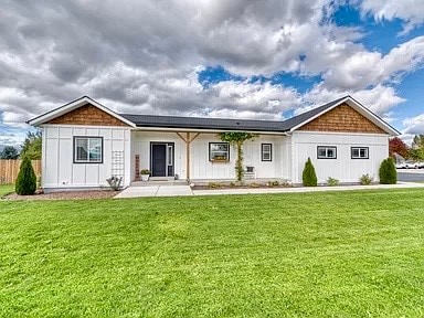The exterior of the house features a blend of horizontal and vertical siding, with a gabled roof adorned with natural wood accents. A well-maintained lawn surrounds the front, complemented by neatly clipped shrubs and seasonal flowers. Expansive clouds drift across a blue sky.