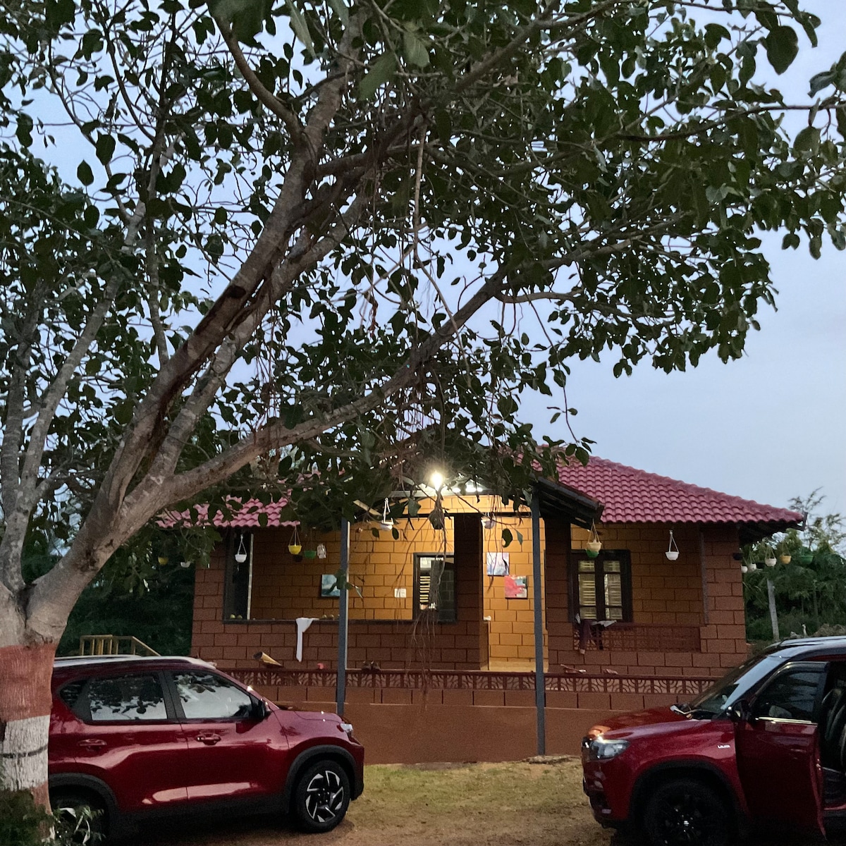 A charming tiny house is set amidst lush greenery, featuring a warm brick exterior and a sloping red roof. The presence of several windows allows natural light to fill the interior, while a front porch is visible, surrounded by trees and parked cars.