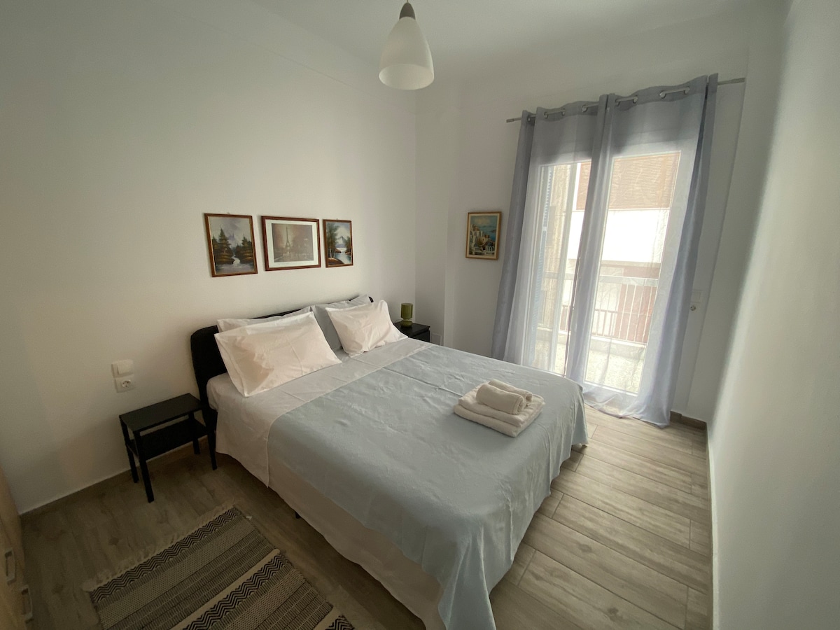 A bright bedroom features a spacious bed with soft linens, flanked by two paintings on the wall. A small nightstand stands beside the bed. Sheer curtains frame a window, allowing natural light to fill the room, while a light fixture hangs above.