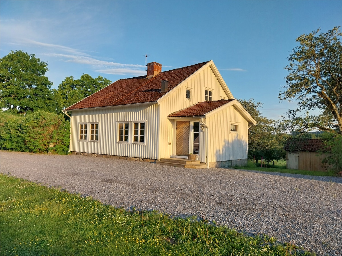 A charming white wooden house features a sloped roof with red tiles, complemented by multiple windows. A gravel driveway leads to the entrance, surrounded by lush greenery and trees. The serene rural setting provides a peaceful atmosphere.