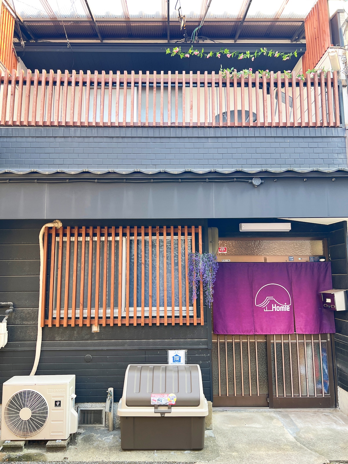 The exterior of a two-story traditional Japanese house features wooden slats and a purple curtain with the word 'Homie.' An air conditioning unit is positioned beside a storage box. A small vine is observed above the entrance, enhancing the welcoming appearance.
