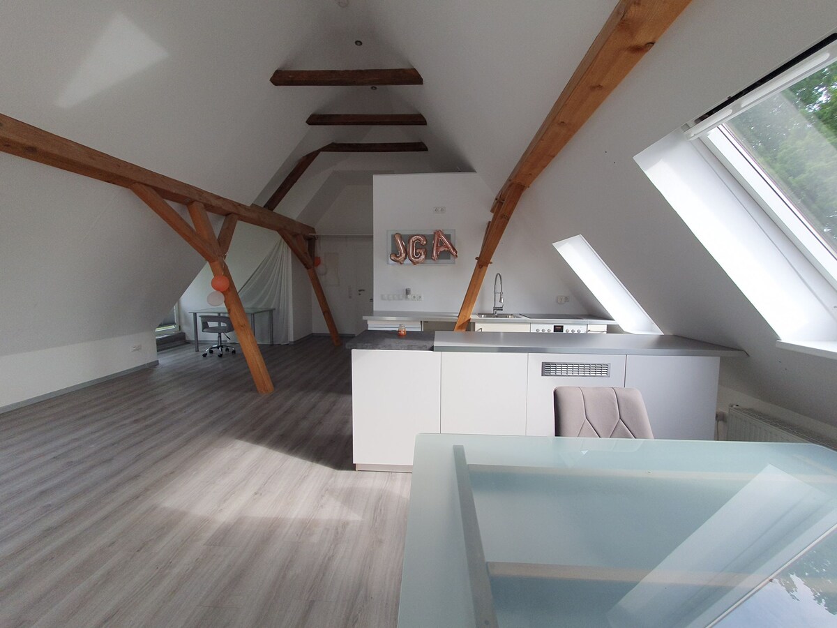 An open living area showcases high vaulted ceilings supported by wooden beams. A modern kitchen is equipped with minimalist cabinetry, while a glass dining table is positioned near a large window that allows natural light to fill the space.