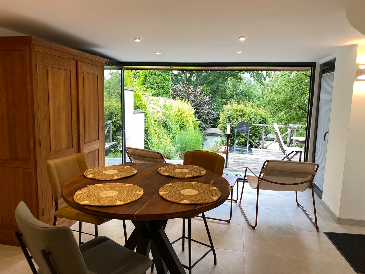 A dining area is visible with a round wooden table set for four, featuring woven placemats. Surrounding chairs offer seating comfort. A large glass door opens to a lush garden, enhancing the indoor-outdoor connection. Natural light brightens the space.