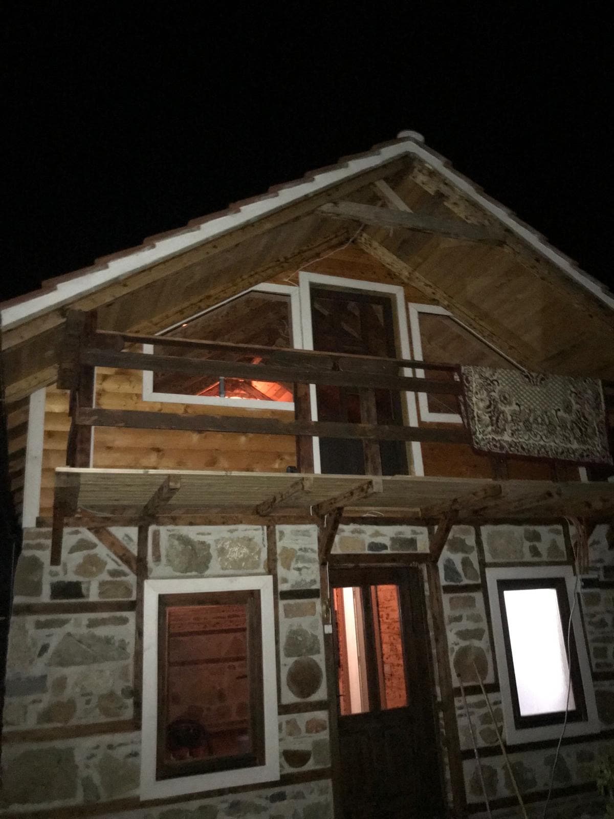 A charming wooden house is illuminated by warm light from the windows, showcasing its rustic stone exterior. The balcony above features wooden beams and decorative elements, while the door at the entrance invites guests inside. The roof is adorned with traditional tiles.