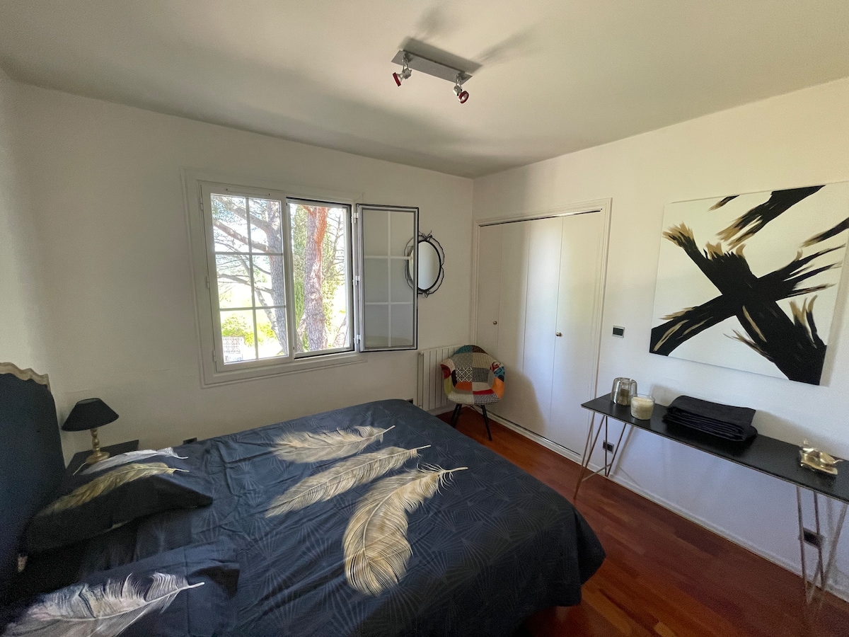 A well-decorated bedroom features a spacious bed with dark bedding adorned with feather patterns. A large window allows natural light to fill the room, complemented by a small chair in the corner and a minimalist desk positioned nearby.