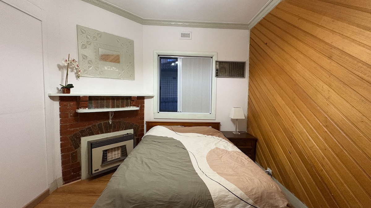 A room featuring a spacious bed with a patterned duvet and a bedside table with a lamp. A brick fireplace adds character, and a large window provides natural light, partially covered with light blinds. The angled wooden wall complements the room's design.