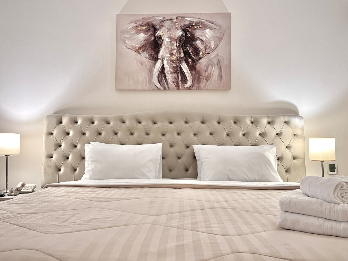 A spacious bed is featured with a neutral-colored quilt and plush pillows. A light-colored upholstered headboard adds elegance, complemented by bedside lamps. Above the bed, an artistic elephant painting enhances the room's decor.