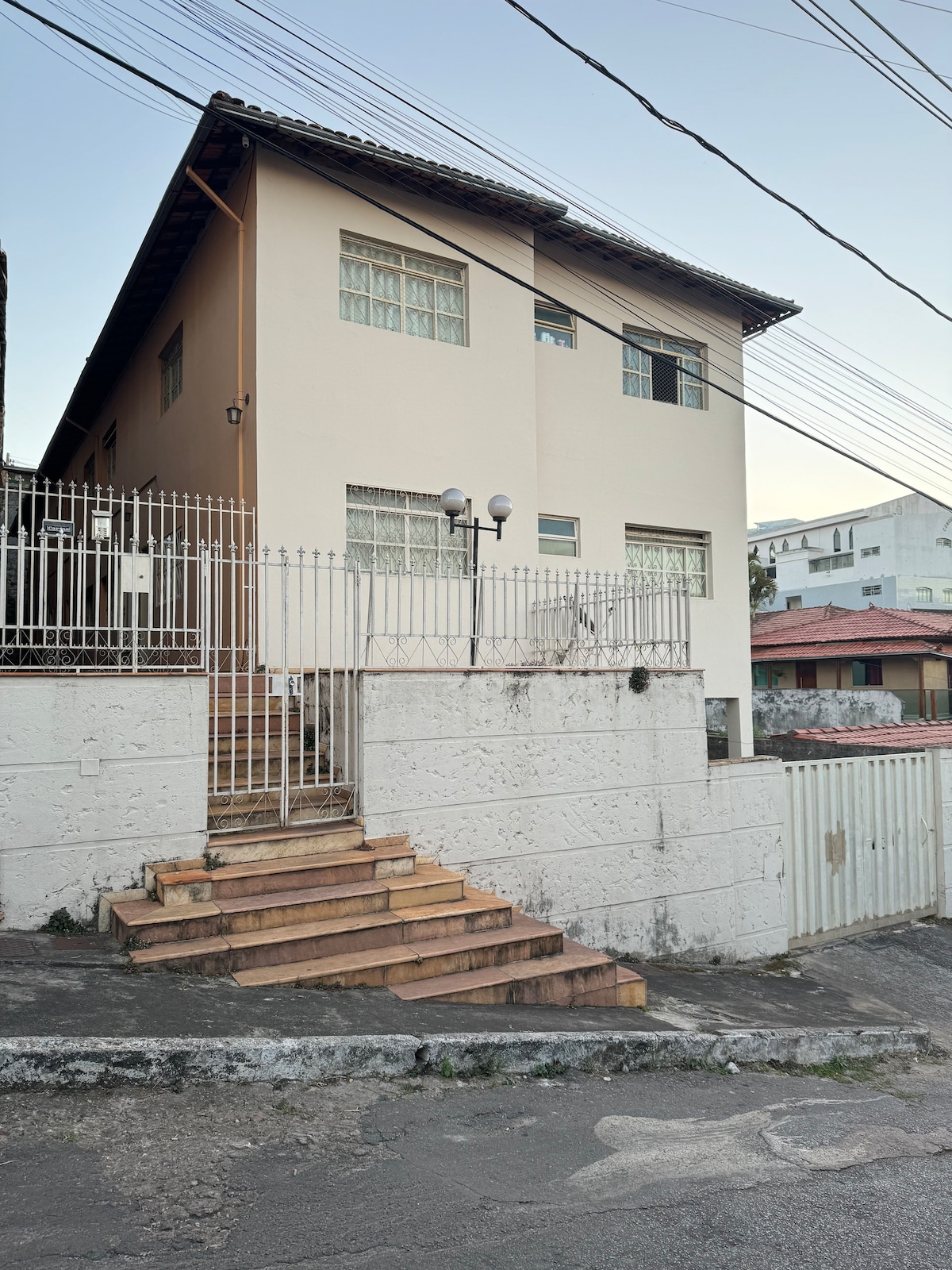 A two-story building is shown, featuring light-colored walls and multiple windows adorned with grilles. Stairs lead up to the entrance, flanked by a low stone wall. The surrounding area is characterized by a mix of residential structures and overhead power lines.
