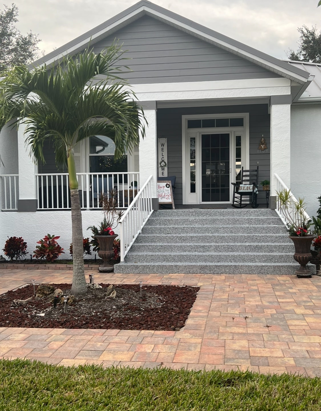 The entrance showcases a charming porch with a rocking chair, flanked by potted plants. Stair steps lead up to a glass front door, while a welcoming sign is displayed nearby. A landscaped area features patterned stonework and a neatly trimmed lawn.