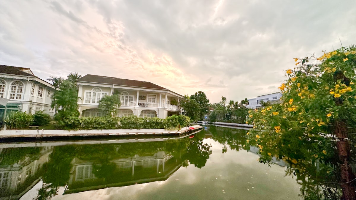 A serene view of the canal reflects the colorful surroundings, framed by lush greenery and blooming yellow flowers. The water's surface mirrors the cloudy sky, while a modest boat is moored along the floating dock, hinting at the village's tranquil lakeside ambiance.