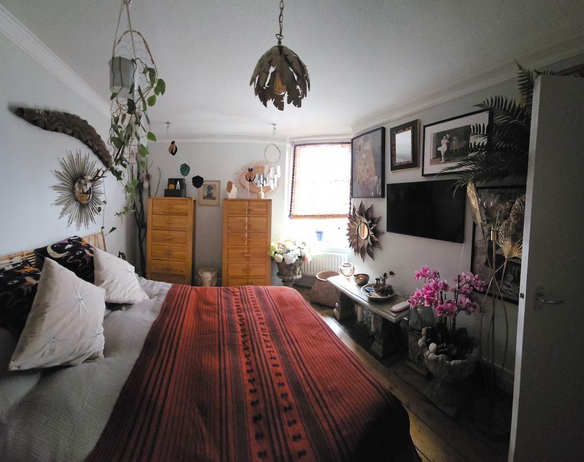 A charming bedroom is presented with a spacious bed adorned with a warm red blanket. Two wooden dressers stand against the wall, while decorative plants and framed artwork add character to the room. A television is mounted opposite the bed, enhancing the cozy atmosphere.