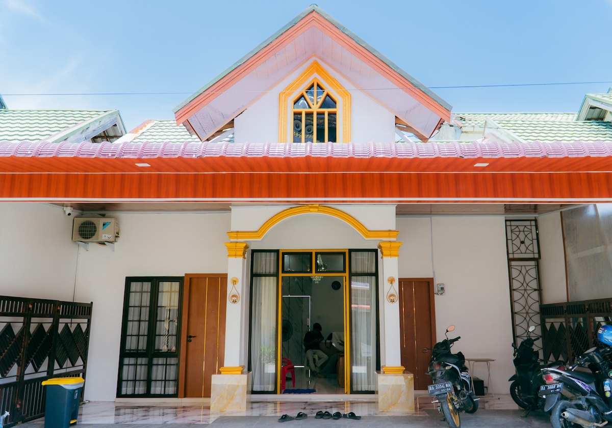 A charming entrance showcases a dual-colored façade with an arched doorway framed by elegant details. The porch is open and features a refreshing layout with visible chairs and a pathway. Two motorcycles are parked nearby, enhancing the accessible atmosphere of the home.