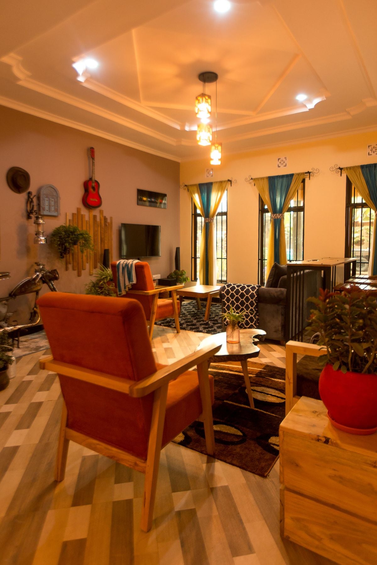 A warm living room is presented with a mix of seating options, including a wooden chair and a stylish couch. Bright blue curtains frame large windows, while decorative elements like a guitar and plants add character. Soft lighting enhances the inviting ambiance.