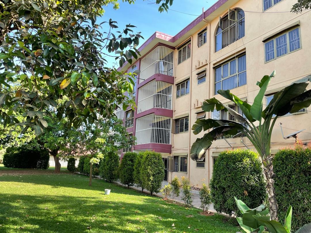 A three-story building with large windows is surrounded by a well-maintained green lawn and lush trees. The façade features a mix of beige and burgundy accents, providing a harmonious blend with the landscape. Shrubs line the pathways leading to the entrance.