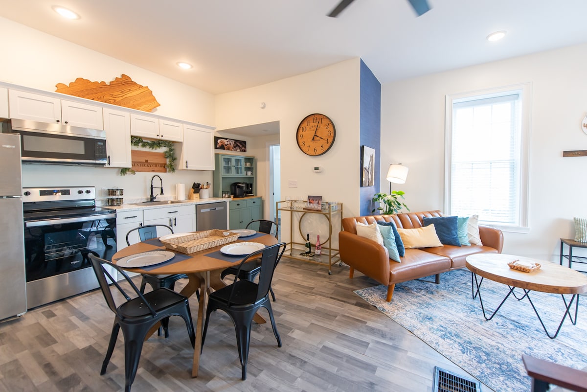 A modern kitchen is featured with stainless steel appliances and white cabinetry, complemented by a round dining table set for four. A cozy living area contains a brown leather sofa and two circular coffee tables, with light filtering through a large window.