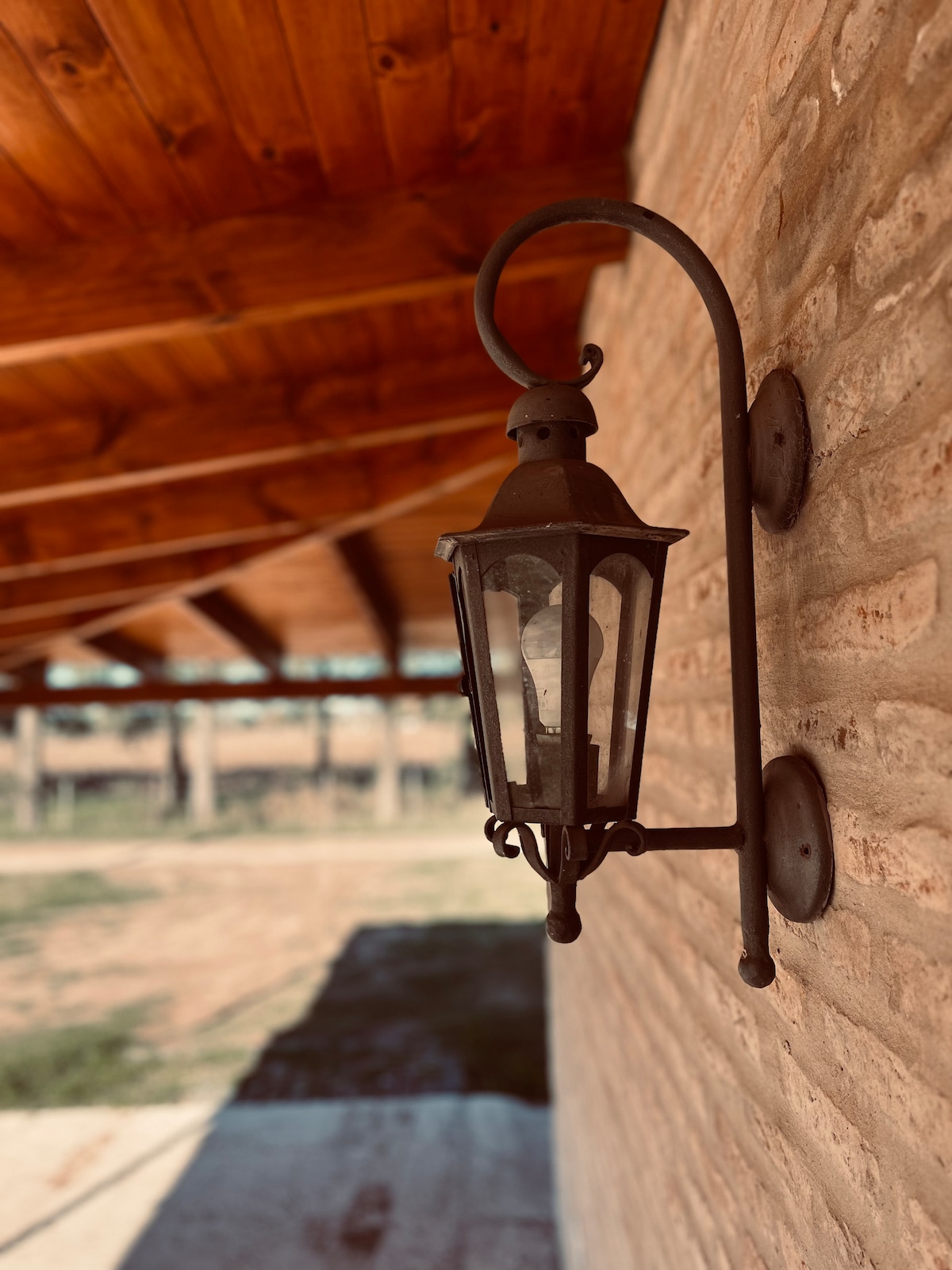 A classic lantern is mounted on a rustic stone wall, providing a charming accent. The lantern features a curved arm and a glass enclosure, while the surrounding area is shaded by a wooden roof structure, enhancing the serene outdoor setting.