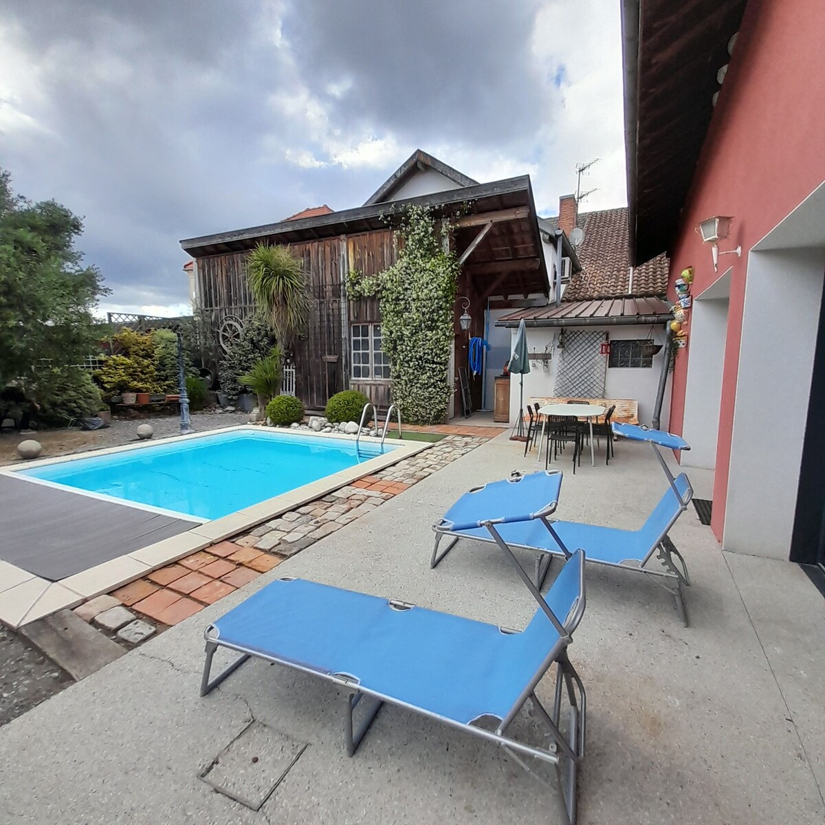 An outdoor area features a rectangular swimming pool surrounded by a stone and tiled patio. Two blue lounge chairs are positioned for relaxation, while a dining table with chairs is ready for outdoor meals. Lush greenery and wooden accents create a serene atmosphere.