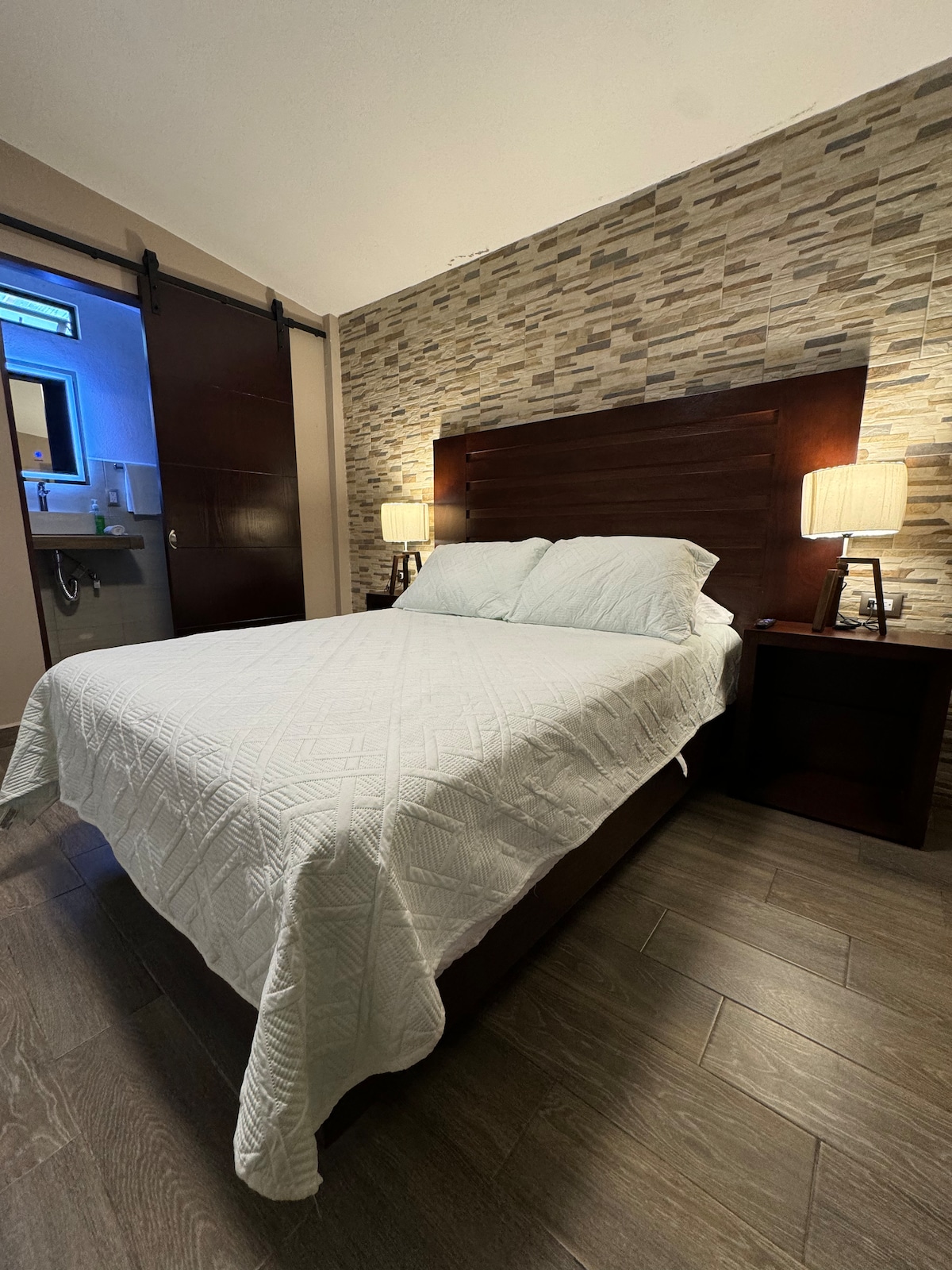 A cozy room features a dark wood bed with a white quilted cover. On either side, matching bedside tables with lamps are positioned. A textured stone wall provides a modern backdrop, while a bathroom area can be seen in the reflection of a nearby glass door.