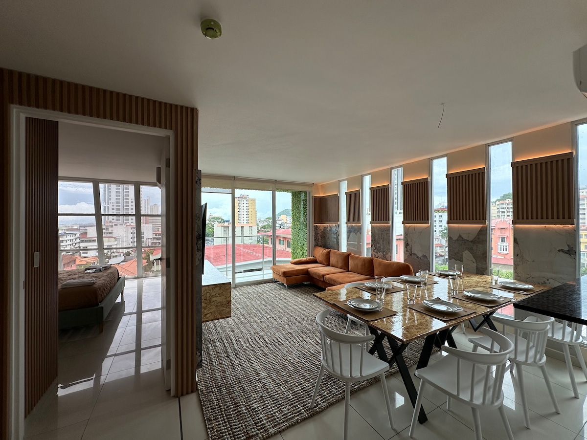 An open living area features a modern dining table with place settings, complemented by a spacious, L-shaped sofa. Large windows provide ample natural light, with a view of the balcony beyond. A separate sleeping area can be seen in the background.
