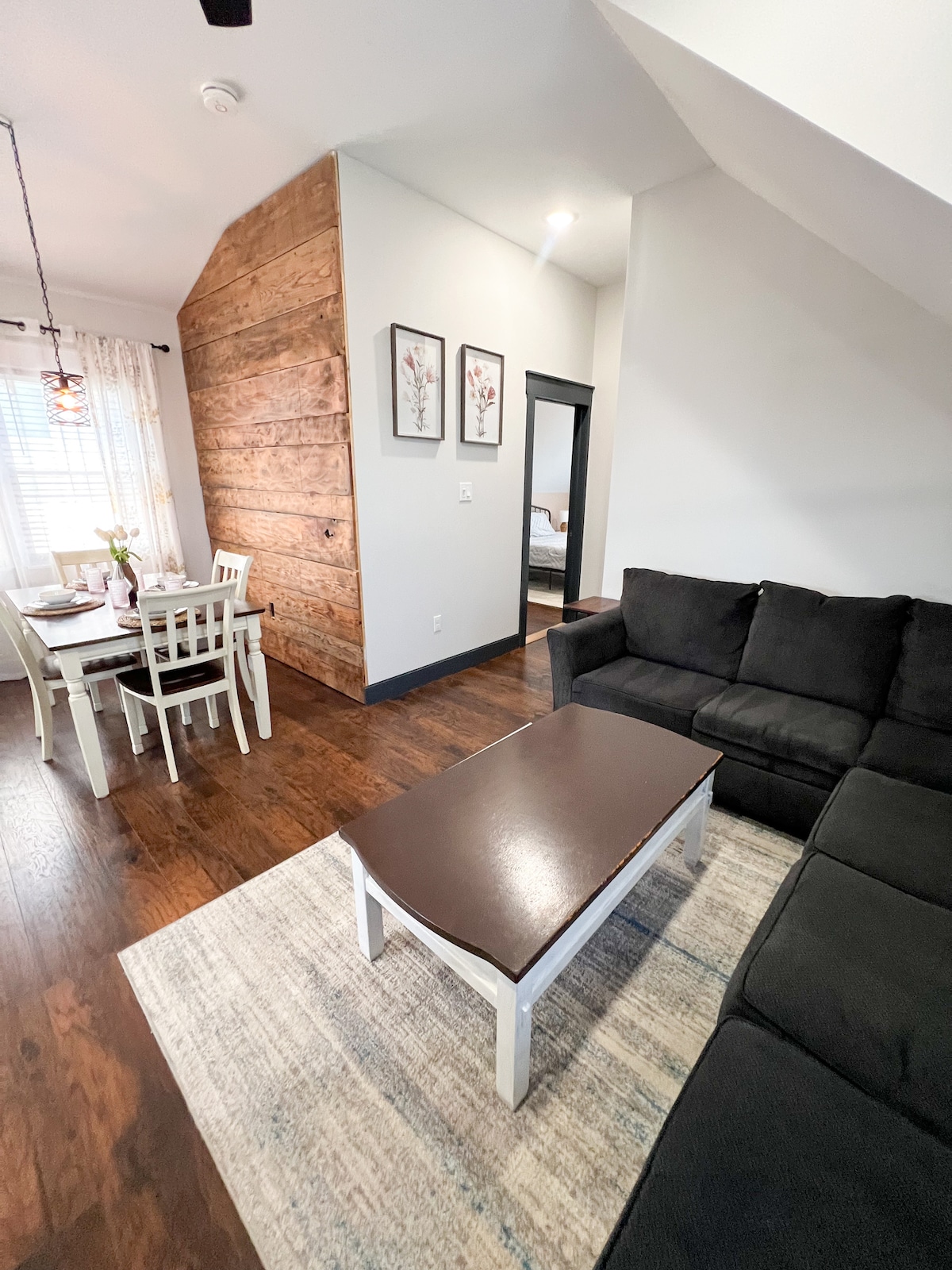 A modern living space features a dark sectional sofa positioned beside a coffee table. A dining area with a light-colored table and chairs is visible, complemented by wood accents on the walls. Natural light filters through sheer curtains, enhancing the warm atmosphere.