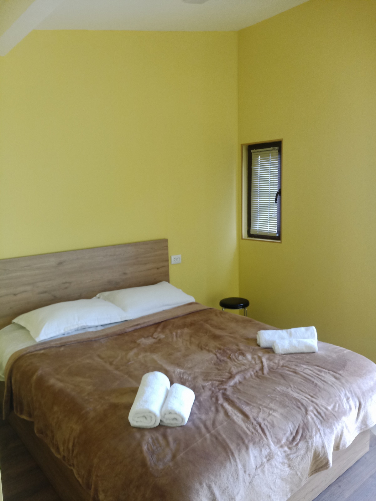 A sleeping area features a spacious bed dressed in a brown blanket, complemented by white linens. Two rolled towels are neatly arranged on the bed. A small stool is positioned nearby, next to a window with horizontal blinds allowing gentle light to filter in.