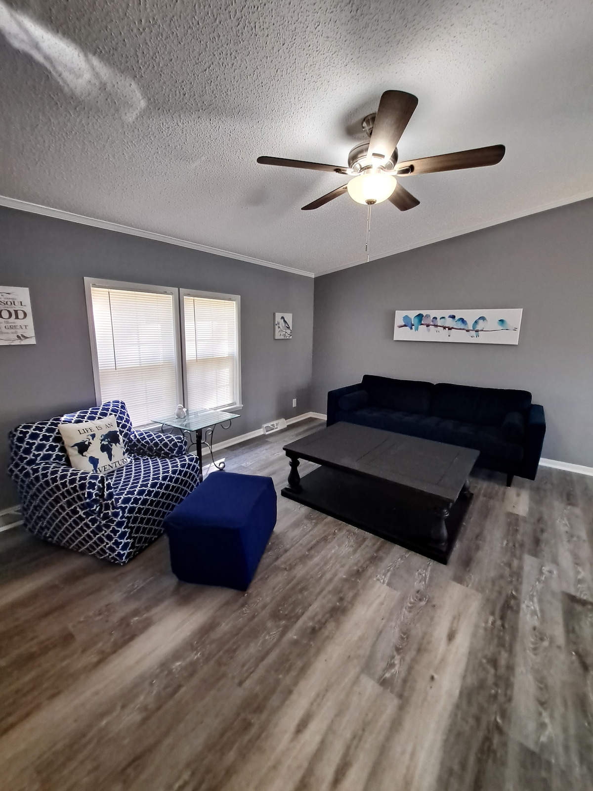The living area is brightened by natural light filtering through two windows. A dark coffee table is positioned at the center, accompanied by a navy blue sofa and an upholstered chair with a patterned design. A ceiling fan adds a functional touch to the space.