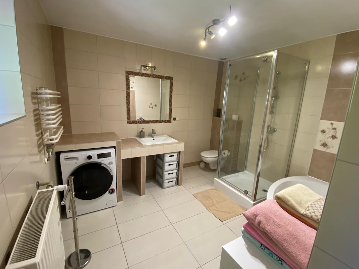 A modern bathroom features light neutral tiles and a glass shower enclosure. A washing machine is positioned near the vanity, which includes a sink and storage drawers. A large mirror is mounted above the sink, complemented by soft lighting. Towels are neatly arranged on a shelf.