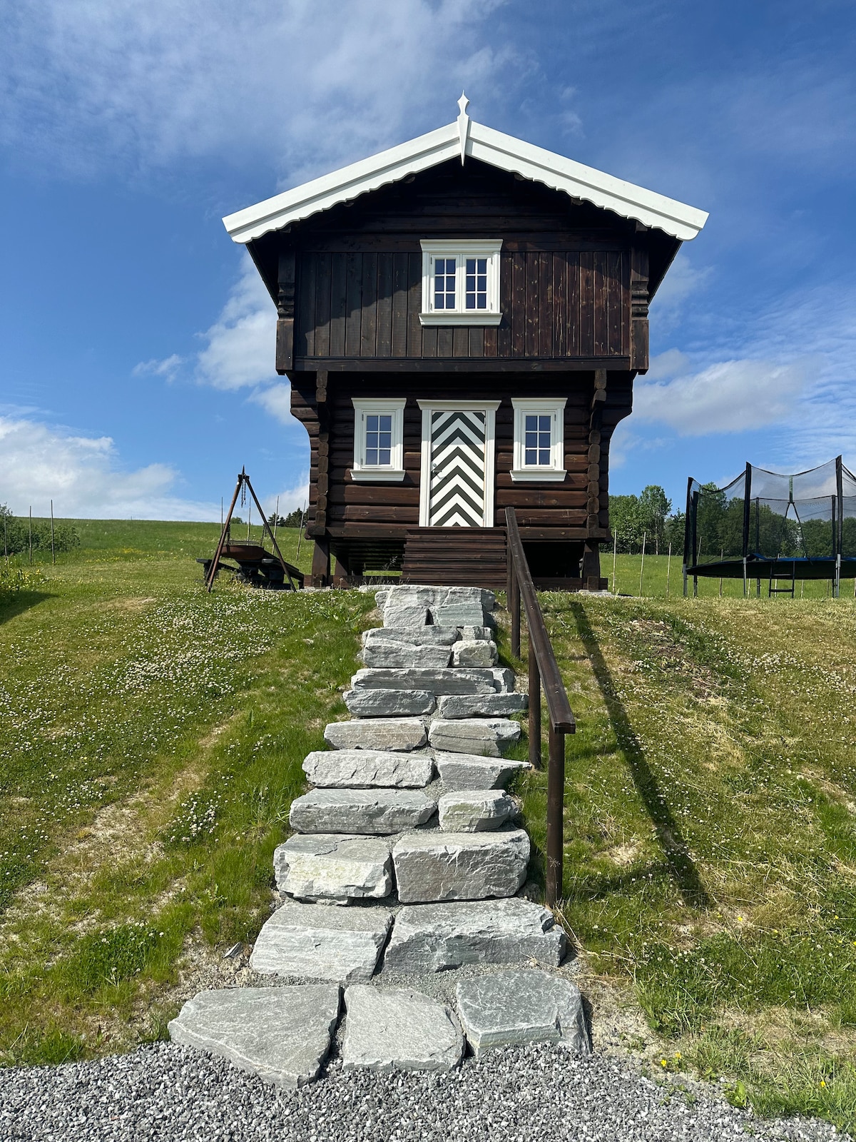 A traditional wooden building sits prominently against the blue sky. Stone steps lead up to the entrance, framed by a grassy area. A swing set can be seen to one side, with a trampoline situated in the background, enhancing the outdoor space.