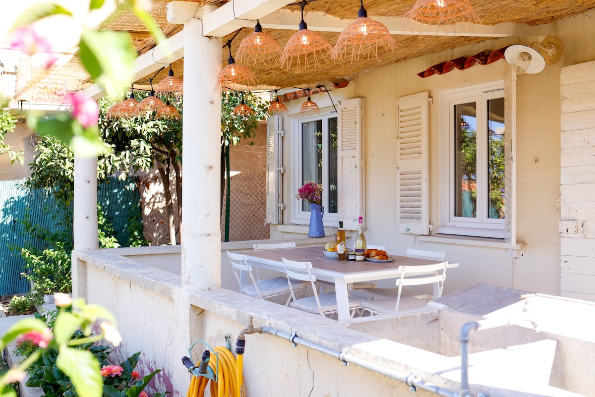 An outdoor terrace features a spacious dining table set for six, surrounded by natural light filtering through decorative pendant lights. Fresh flowers in a vase add a touch of charm, while the walls display a neutral color palette, creating an inviting atmosphere.