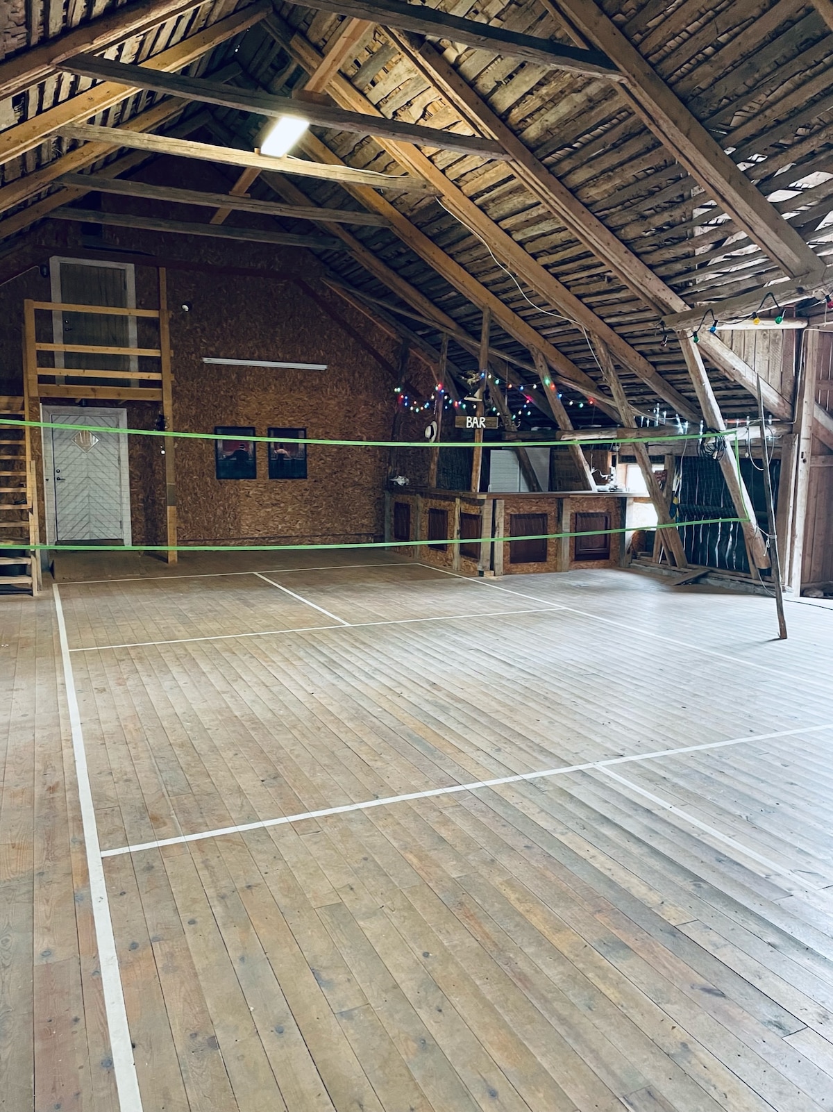 An expansive indoor space features a wooden floor and high ceilings supported by exposed beams. Volleyball netting is set up in the center, while colorful lights are strung along the walls. Stairs lead to a loft area, providing additional space.