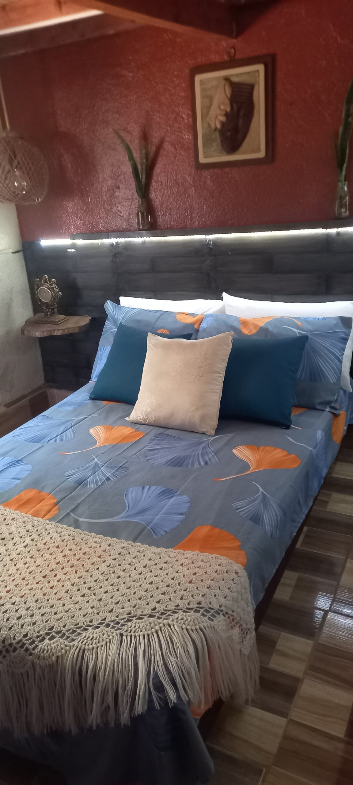 A cozy sleeping area features a bed adorned with a colorful floral-patterned blanket and several decorative pillows. A bedside table displays a small decorative item, and the room is accentuated by textured walls and subtle lighting.