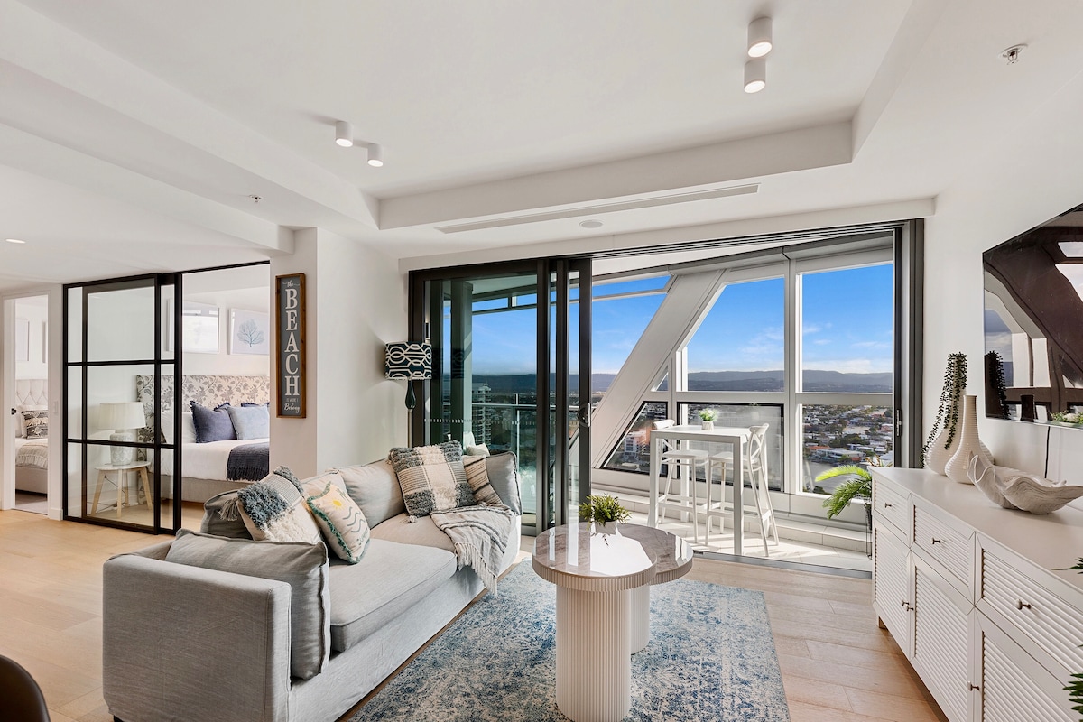 A bright, open living area features a contemporary sofa adorned with decorative pillows. A round coffee table sits on a soft area rug, and large glass doors provide stunning views of the landscape outside, while the adjoining glass-walled bedroom is visible in the background.