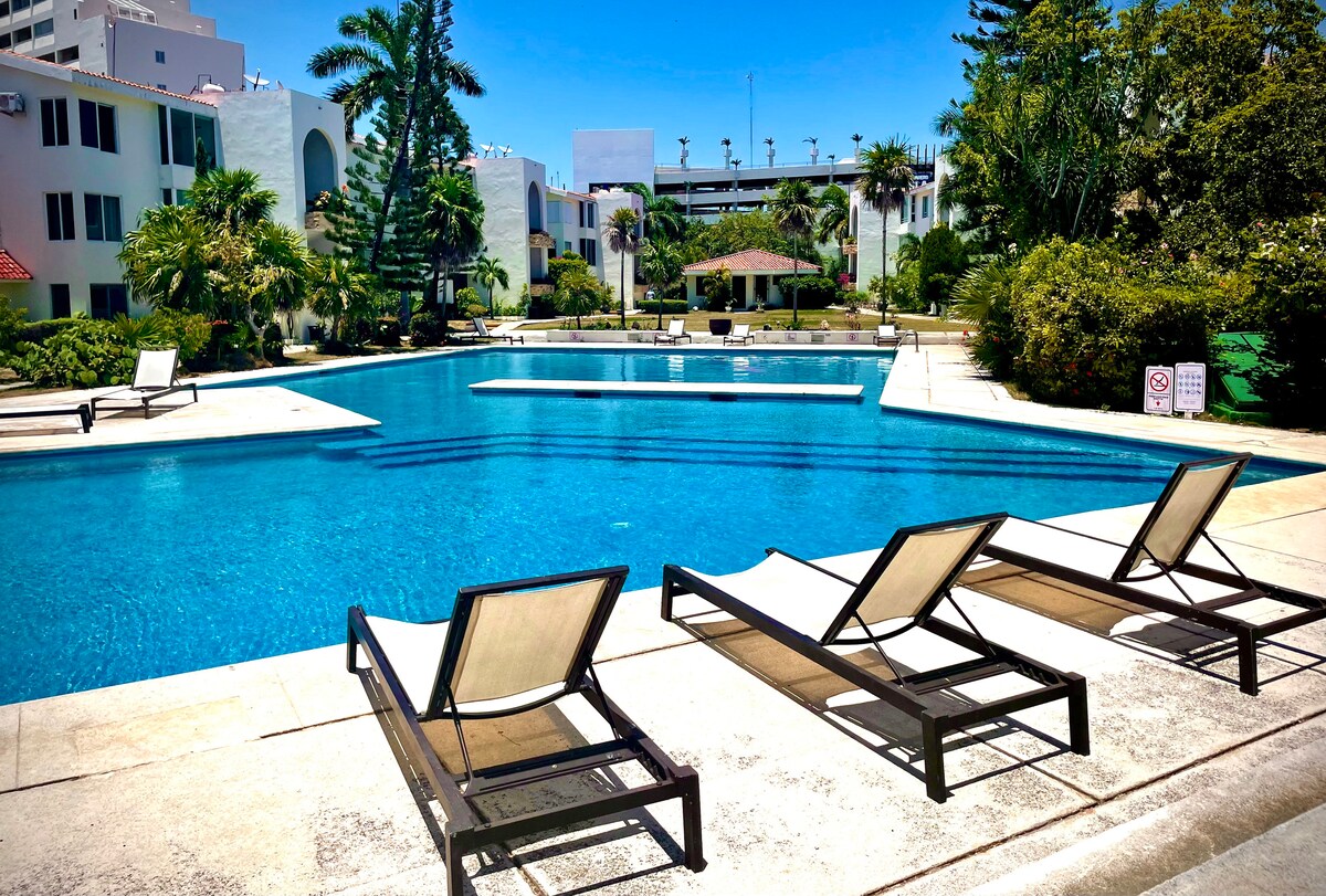 A well-maintained swimming pool is surrounded by lush landscaping, with multiple lounge chairs positioned along the deck. An inviting palapa provides shade, while the clear water reflects the blue sky above. The area offers a serene escape for relaxation and leisure activities.